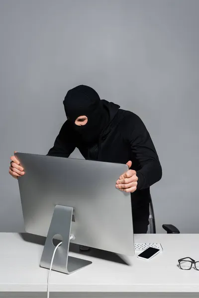 Hacker mit Sturmhaube nimmt Computermonitor in der Nähe von Smartphone isoliert auf grau — Stockfoto