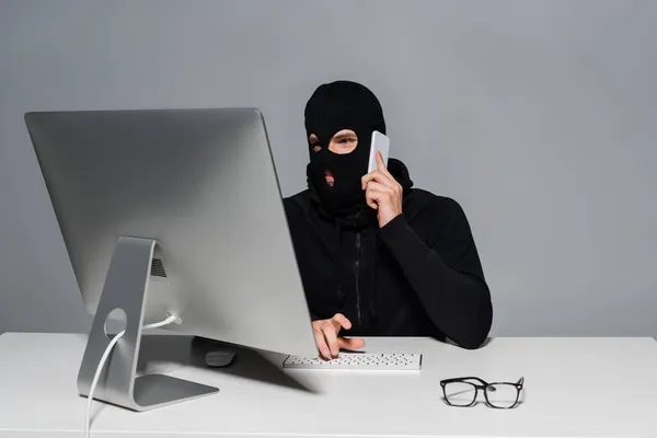 Hacker in balaclava talking on smartphone and using computer near eyeglasses isolated on grey — Stock Photo