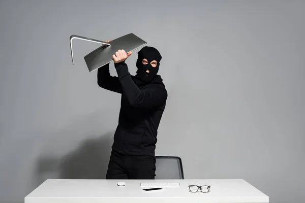 Pirata informático enojado que sostiene el monitor de la computadora cerca del teléfono inteligente y las gafas en la mesa sobre fondo gris - foto de stock