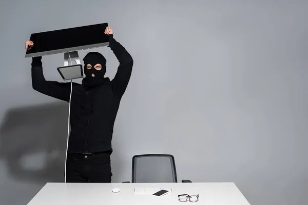 Хакер в Балаклаві тримає монітор комп'ютера біля мобільного телефону на столі на сірому фоні — стокове фото