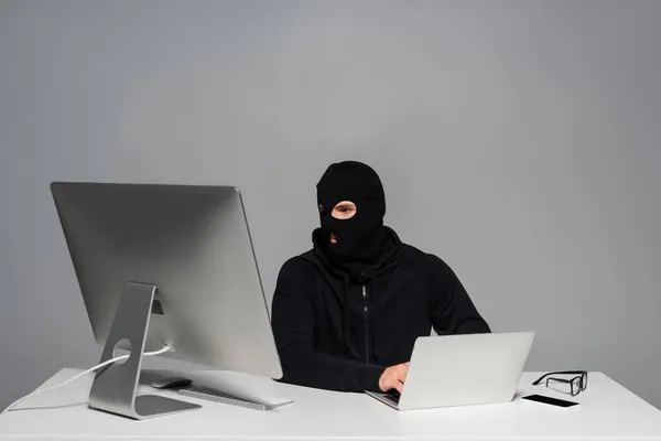 Hacker en pasamontañas con ordenador y portátil cerca de teléfono inteligente en la mesa aislado en gris - foto de stock