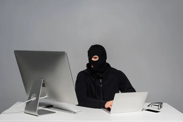 Hacker em balaclava usando computador e laptop perto de smartphone na mesa isolado em cinza — Fotografia de Stock