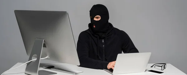 Hacker in passamontagna utilizzando laptop vicino al computer e smartphone isolato su grigio, banner — Foto stock