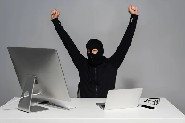 Aufgeregte Hacker mit Sturmhaube zeigen Ja-Geste in der Nähe von Gadgets auf grauem Hintergrund — Stockfoto