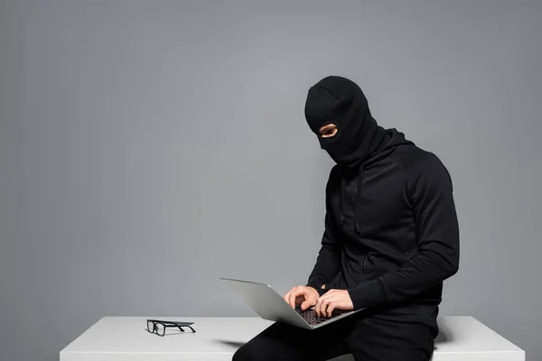 Hacker in balaclava using laptop near eyeglasses on table isolated on grey — Stock Photo