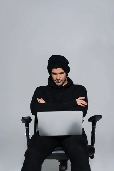 Хакер в балаклаве сидит со скрещенными руками рядом с ноутбуком изолирован на сером — стоковое фото