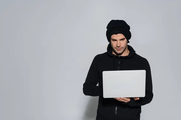 Hacker en pasamontañas usando laptop sobre fondo gris - foto de stock
