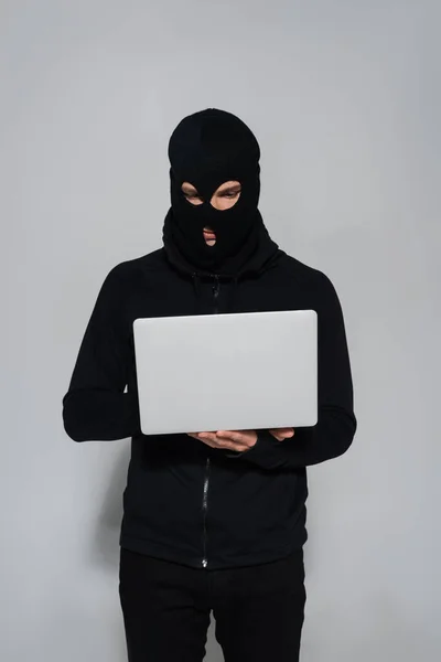 Hacker in black balaclava using laptop on grey background — Stock Photo