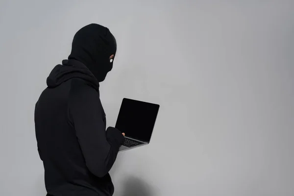 Hacker en pasamontañas usando laptop con pantalla en blanco sobre fondo gris - foto de stock