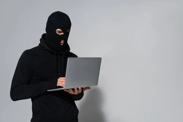 Hacker in balaclava using laptop on grey background — Stock Photo