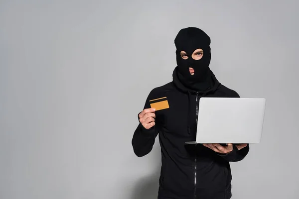 Hacker em balaclava segurando cartão de crédito e laptop em fundo cinza — Fotografia de Stock