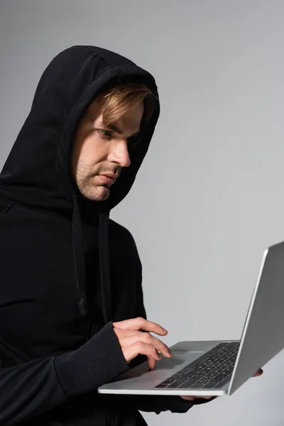 Joven hacker utilizando el ordenador portátil, mientras que de pie aislado en gris - foto de stock