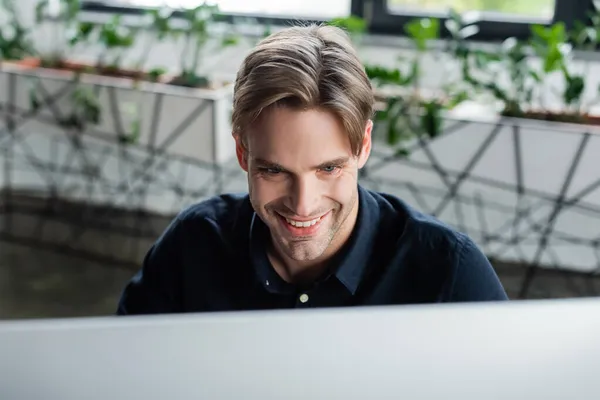 Programador sonriente mirando el monitor borroso de la computadora en la oficina - foto de stock