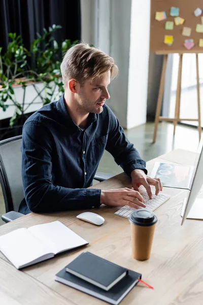 Jeune programmeur utilisant un ordinateur près du café pour aller et les ordinateurs portables dans le bureau — Photo de stock