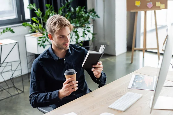 Programmatore che tiene tazza di carta e notebook vicino al computer in ufficio — Foto stock