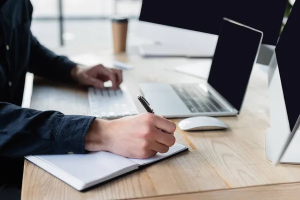 Cropped view of programmer writing on notebook near computers on blurred background — Stock Photo
