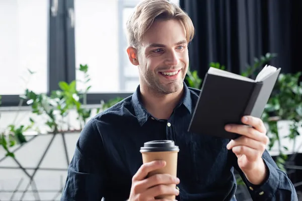 Programador sorridente olhando para notebook e segurando café para ir no escritório — Fotografia de Stock