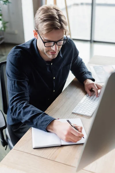 Programmer in eyeglasses writing on notebook and using computer in office — Stock Photo