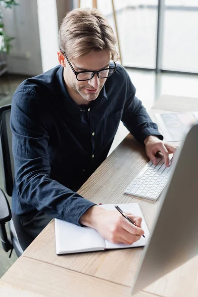 Programmierer mit Brille schreibt auf Notizbuch und benutzt Computer im Büro — Stockfoto