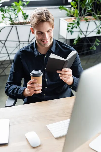 Fröhlicher Programmierer hält Pappbecher und Notizbuch neben Computer im Büro — Stockfoto