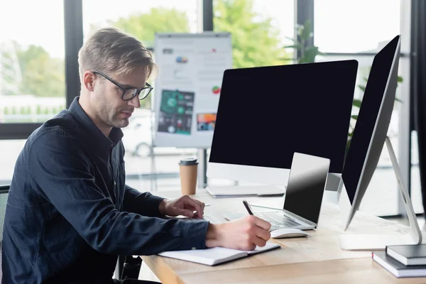 Programmer writing on notebook near computers and coffee in office — Stock Photo