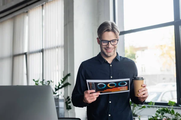 Smiling programmer holding paper and coffee to go near computer in office — Stock Photo