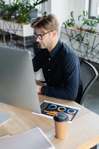 Programmatore in occhiali usando il computer vicino a documento e caffè in ufficio — Foto stock