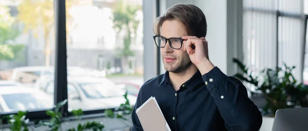 Programmer holding eyeglasses and paper folder near computer monitor in office, banner — Stock Photo