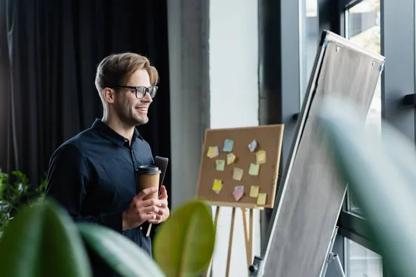 Positiver Programmierer mit Kaffee- und Papiermappe beim Blick auf Flipchart im Büro — Stockfoto