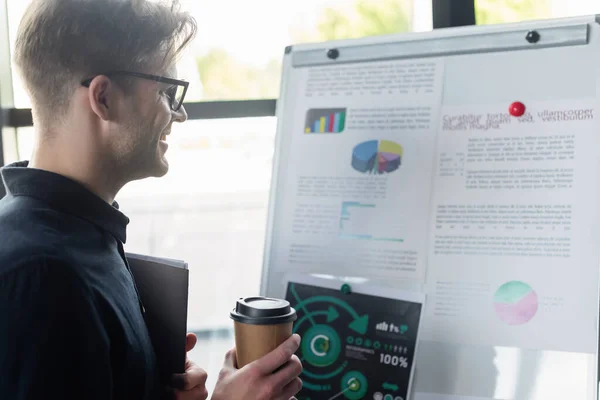 Side view of cheerful programmer with coffee and paper folder looking at charts on flip chart in office — Stock Photo