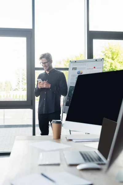 Programador positivo usando smartphone perto de flip chart e computadores no escritório — Fotografia de Stock