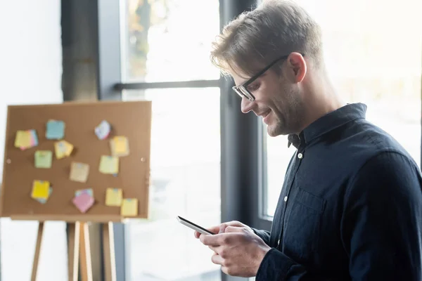 Side view of smiling programmer using smartphone in office — Stock Photo