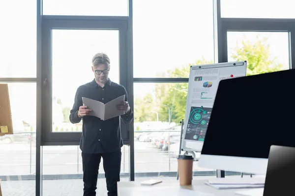 Programador em óculos segurando pasta de papel perto de flip chart e dispositivos no escritório — Fotografia de Stock