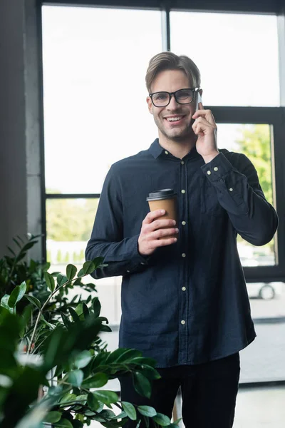 Programador feliz en anteojos hablando en smartphone y sosteniendo taza de papel cerca de las plantas - foto de stock