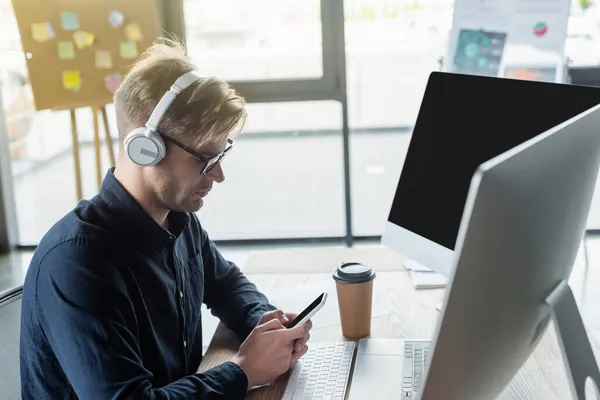 Side view of developer in headphones using smartphone near coffee and computers in office — Stock Photo
