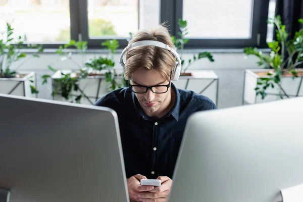 Young programmer in headphones using smartphone near computer monitors — Stock Photo