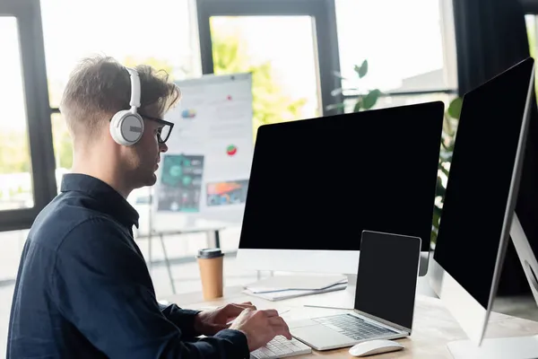 Vista laterale del programmatore in cuffia utilizzando computer vicino alla tazza di carta sul tavolo — Foto stock