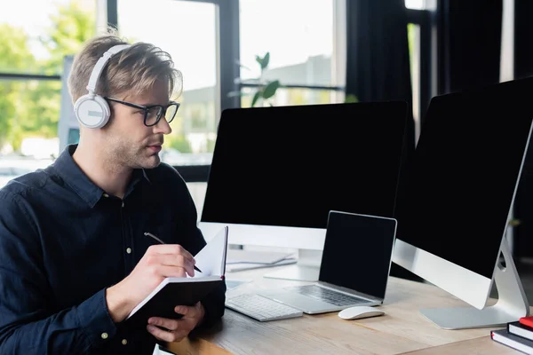 Programmer in headphones holding notebook near computers in office — Stock Photo