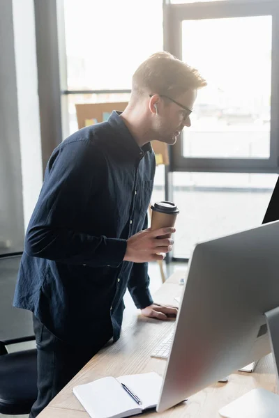 Side view of programmer in earphone holding paper cup near computers and notebook in office — Stock Photo