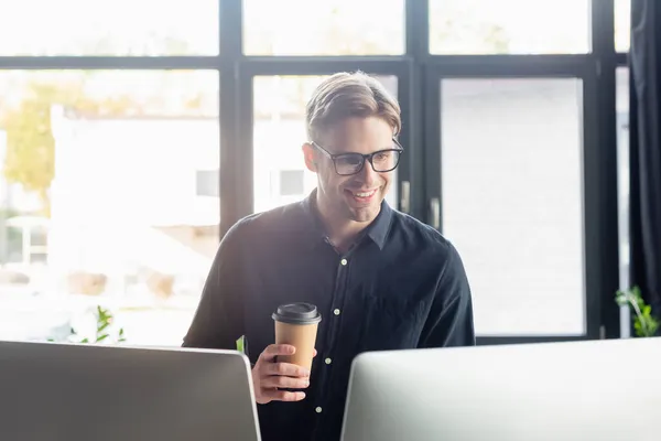 Programador sorridente com café para ir olhar para monitores de computador no escritório — Fotografia de Stock