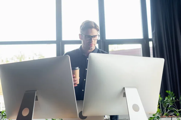 Young programmer in eyeglasses looking at computers and holding coffee to go in office — Stock Photo