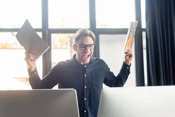 Aufgeregter Programmierer hält Papiermappe und Dokument in der Nähe von Computern im Büro — Stockfoto