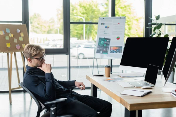 Programador pensativo mirando las computadoras cerca de teléfono inteligente y café en la mesa - foto de stock