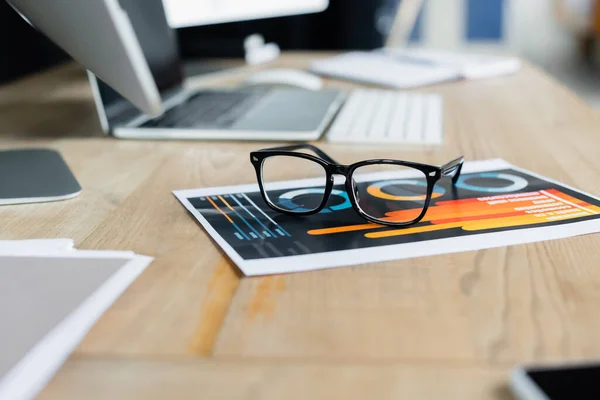 Gafas graduadas en papel con gráficos cerca de computadoras borrosas en la oficina - foto de stock