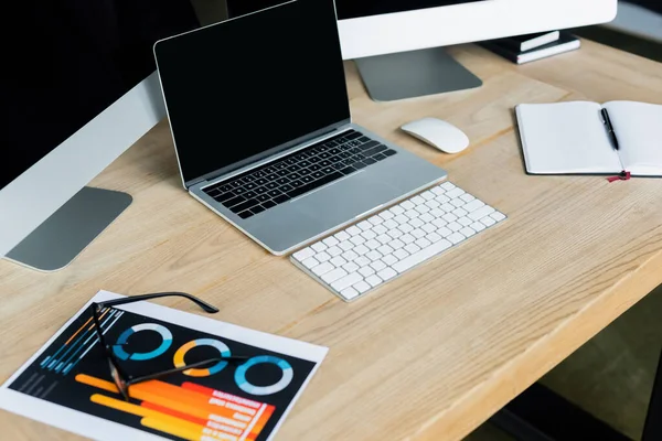 Computadoras con pantalla en blanco cerca de anteojos y papel con gráficos en la mesa - foto de stock