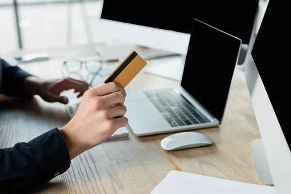 Vista recortada del programador que sostiene la tarjeta de crédito cerca de computadoras con la pantalla en blanco en oficina - foto de stock