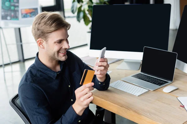 Desenvolvedor positivo usando smartphone e cartão de crédito perto de computadores no escritório — Fotografia de Stock