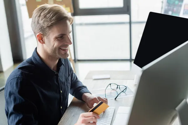 Cheerful programmer holding credit card while using computers in office — Stock Photo