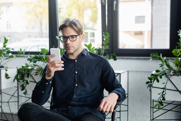 Young developer in eyeglasses using smartphone while sitting on chair in office — Stock Photo