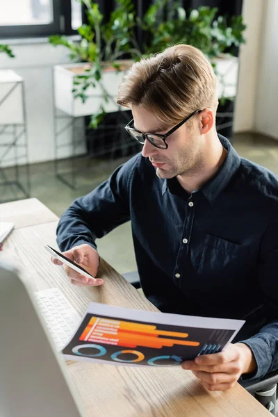 Joven programador usando smartphone y sosteniendo documento cerca de la computadora en la oficina - foto de stock